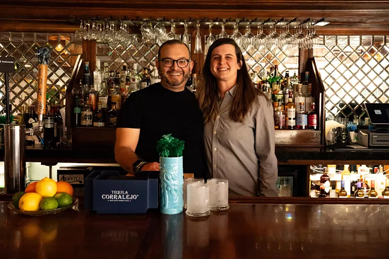 Image: A man in glasses and a black shirt next to a woman in a grey button-down shirt standing behind a bar