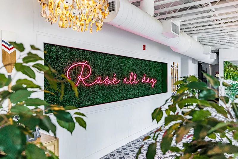 Image: a pink "Rosé All Day" neon sign on a plant-covered background