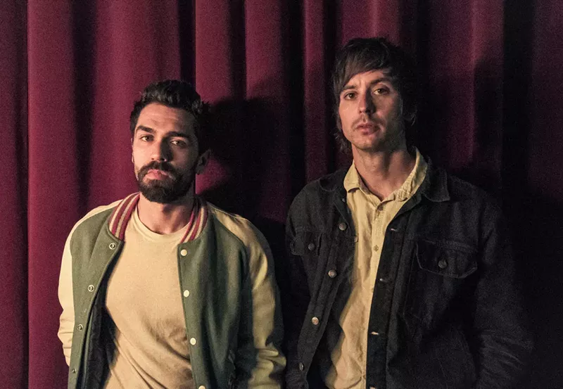 Image: two men in jackets stand in front of a red velvet curtain.