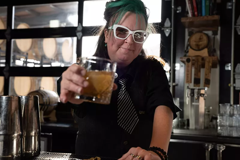 Image: a woman with teal hair and a black and white striped ties holding a cocktail