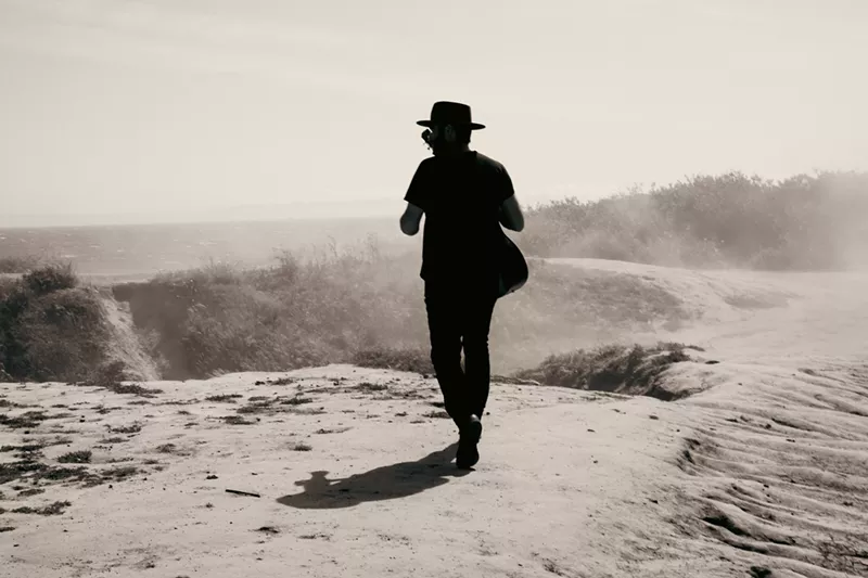 Image: man walking around plains with a guitar strapped on his back