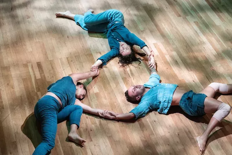 Image: three people wearing blue lie on the floor holding hands