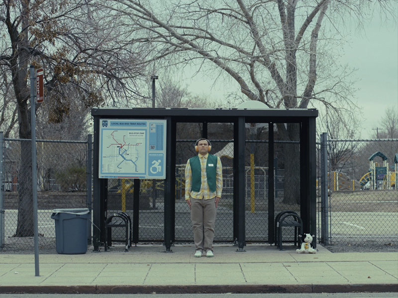 Image: Man with headphones stands at a bus stop.