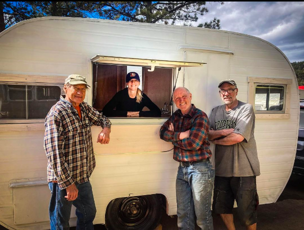 A woman smiles from inside a pop up window in a camper along with three men.