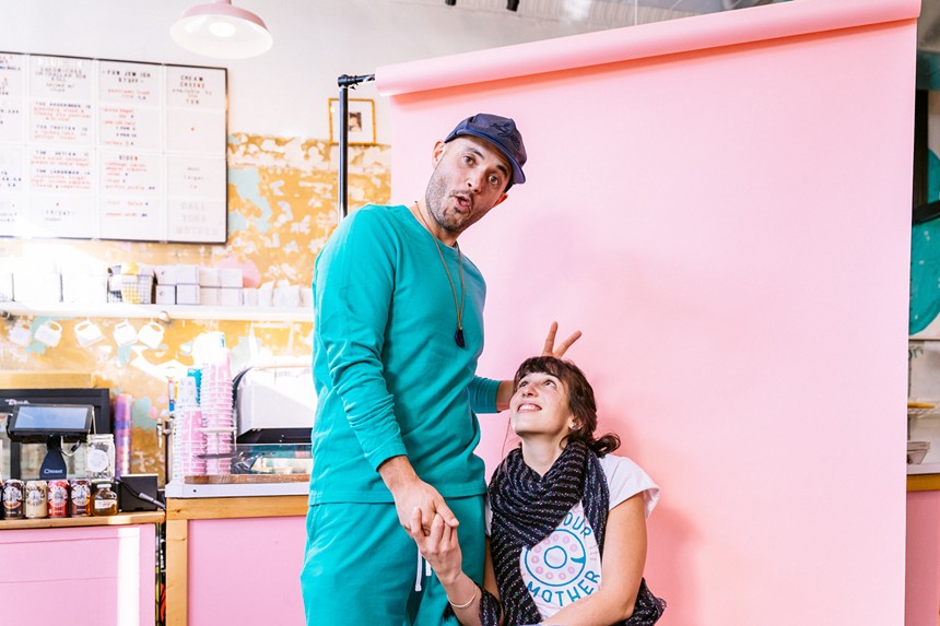 a man in a teal sweatsuit gives bunny ears to a woman sitting down in front of a pink wall