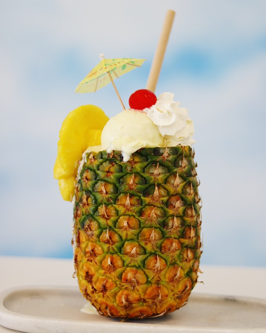A hollowed pineapple filled with smoothie, whipped cream, and a pineapple slice that has been decorated with a paper umbrella and cherry.