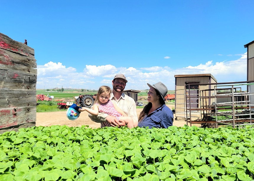 a man holding a young girl standing next to a woman in front of seedlings on a farm