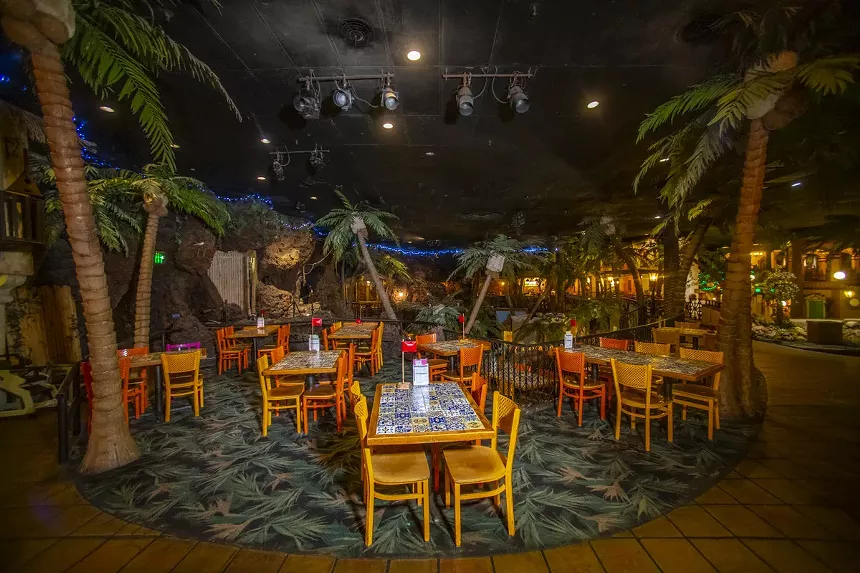 a dining room with tables, chairs and fake palm trees