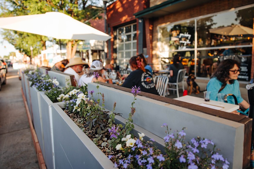 people sitting at tables on a patio lined with flowers in planter boxes