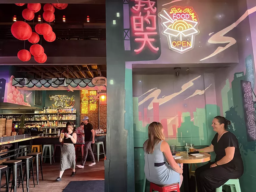 two women sit at a small table under a neon sign inside a restaurant