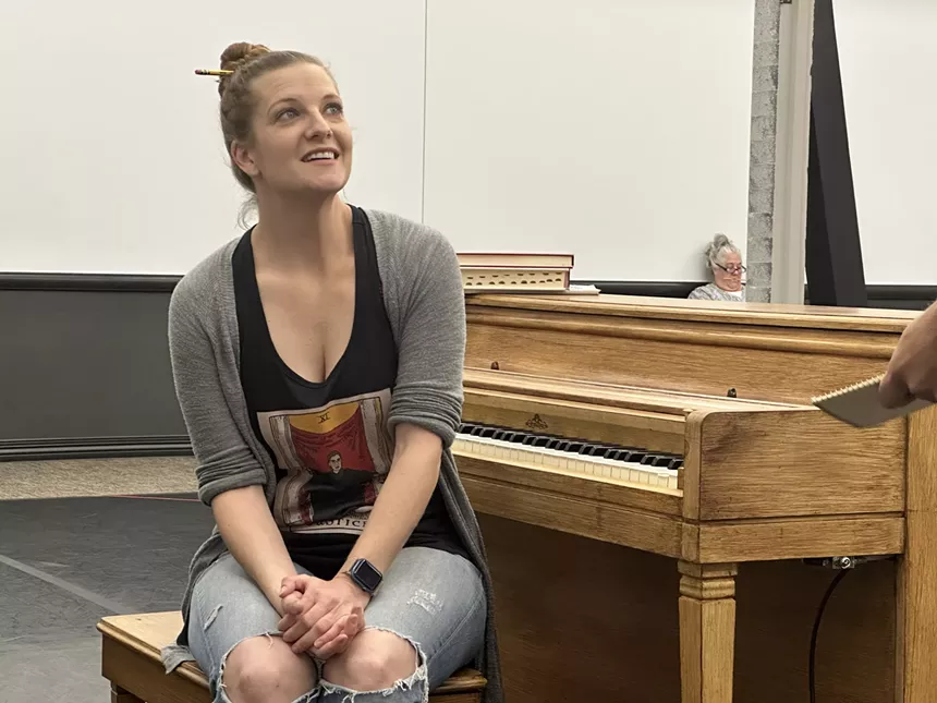 young woman in black tank top and grey sweater sits at a piano