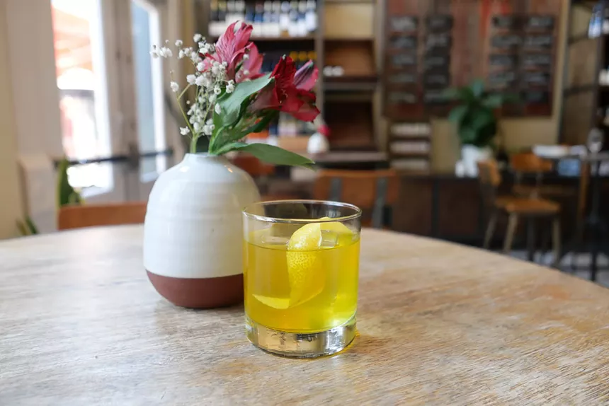 glass of yellow liquid cocktail sits on table next to vase of pink flowers