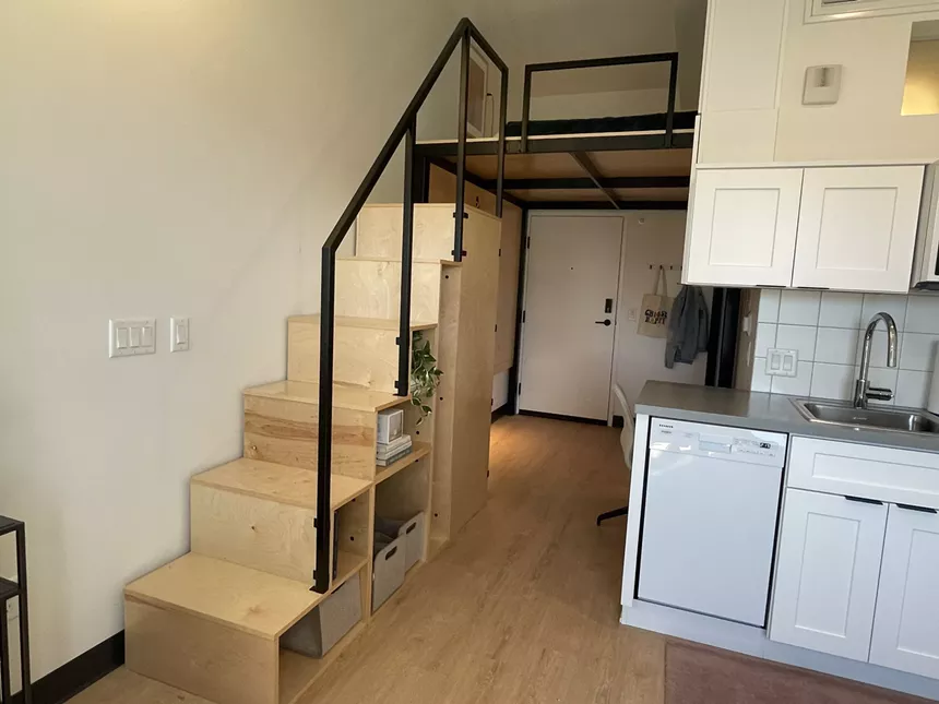 Lightwood stairs with a black metal railing lead up to a space big enough for a mattress.