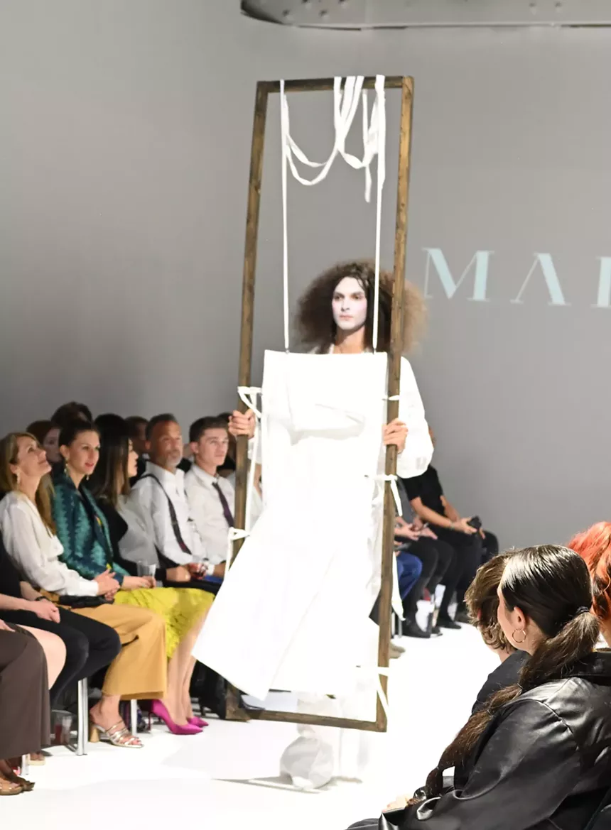 person with face painted white aand wearing white walks down runway holding two sticks with a white sheet of paper inbetween.