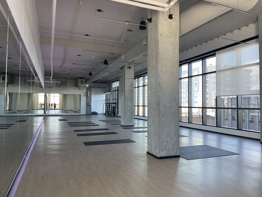 Yoga mats sit atop light wood in a room with exposed concrete beams and floor-to-ceiling windows.