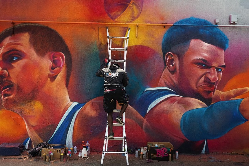 man climbing down ladder while working on a colorful mural of basketball players.