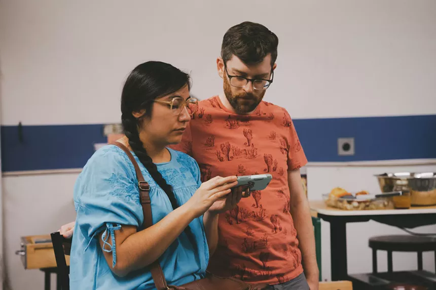 woman in blue shirt, glasses and braid shows phone to man in orange shirt with glasses