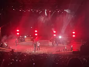 Two men sing on a stage lit in red.
