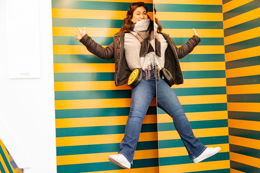 woman in jeans and brown jacket and scarf jumps in green and yellow striped room.