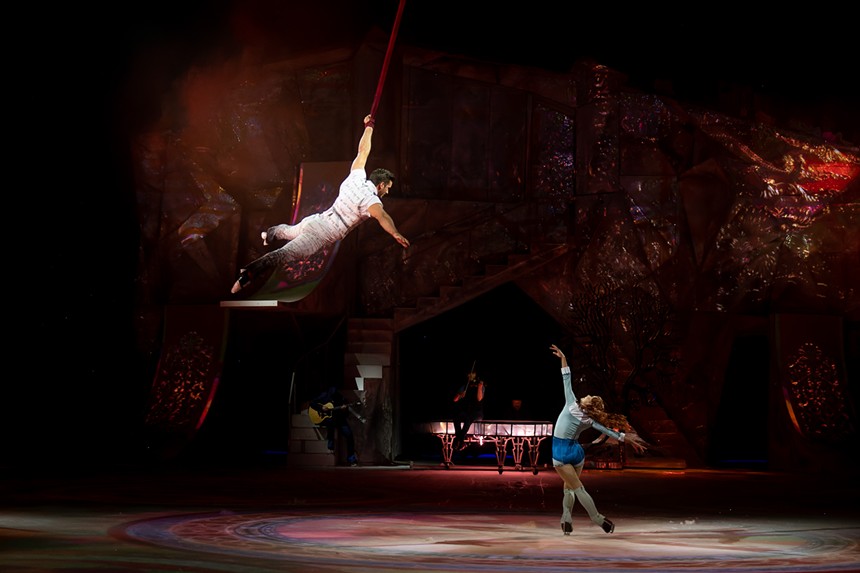 man hanging from an aerial strap while woman skates below on an icerink