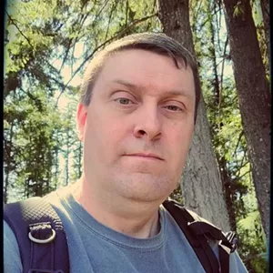 white man looking into the camera while standing in the woods with a backpack and blue shirt