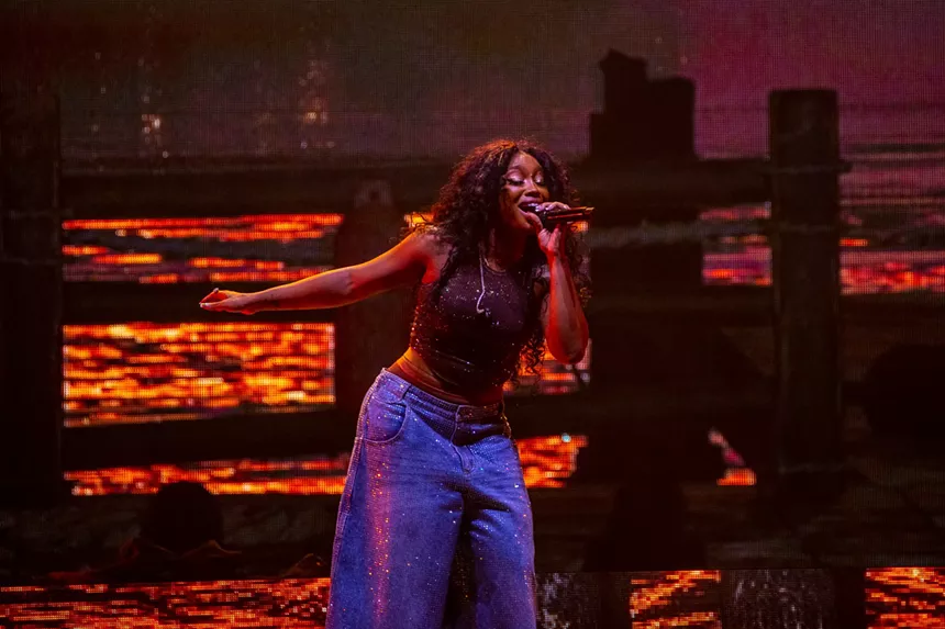 woman in blue pants singing into a mic with an orange background