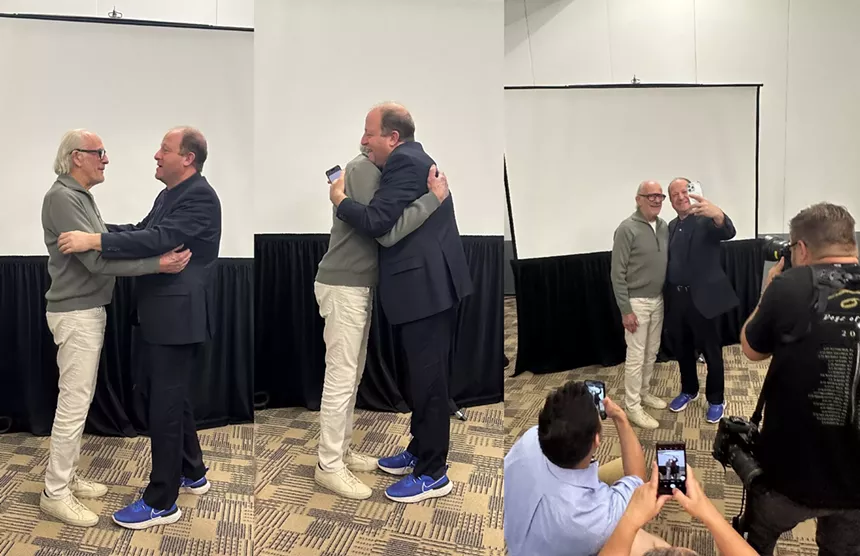 two men hugging at convention in front of mirrors.