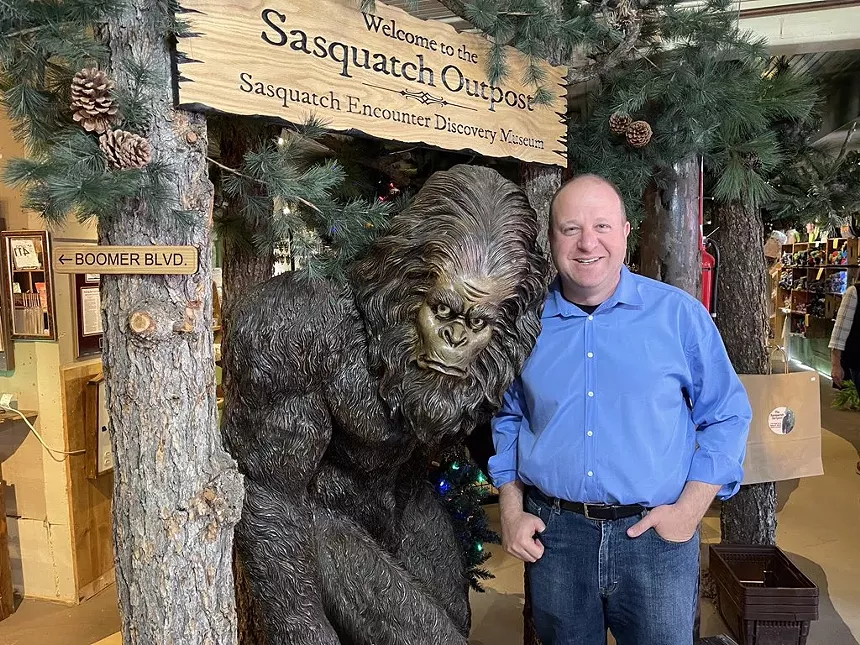 on the left is a model of a bigfoot, on the right is a bald man in a blue shirt posing next to it