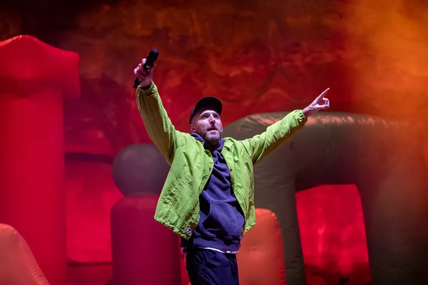 man in neon green jean jacket and baseball hat points both hands in the air