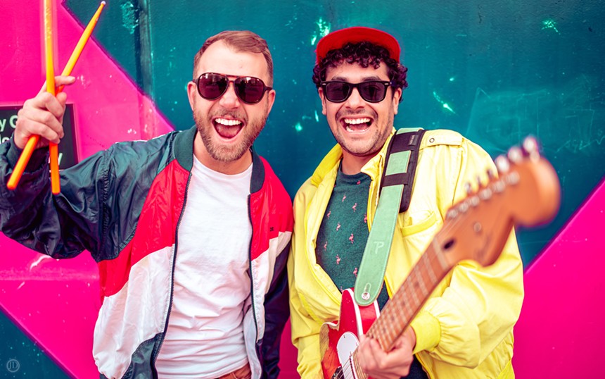 a man holding drumsticks and a man holding a guitar wear sunglasses and smile for a photo.