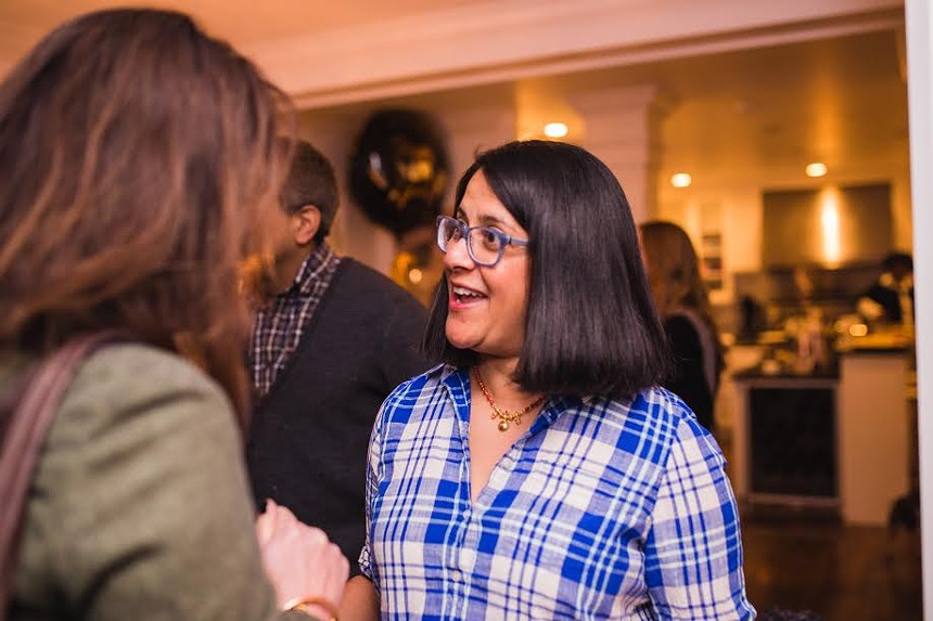 A woman with black hair and glasses in a blue flannel smiles at someone else.