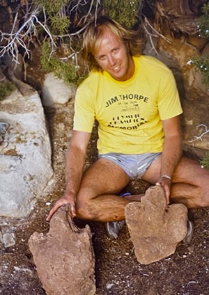 A man in a yellow shirt and jean shorts holds up two red-dirt fossils.