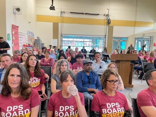 Several members of a crowd at a Denver School Board meeting wear red "Resign DPS" shirts.