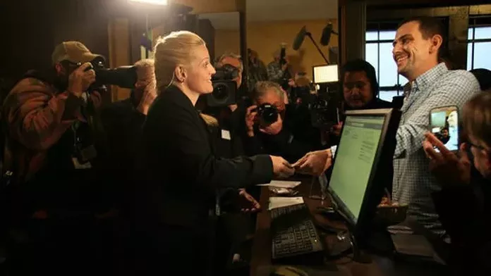 A blonde woman in black sells weed to man as they are surrounded by cameras