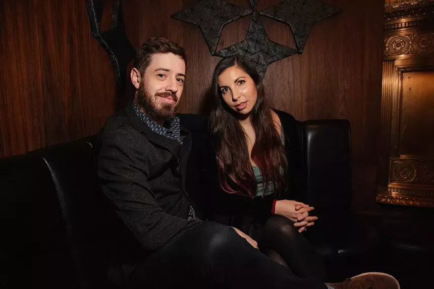 bearded man in black jacket and brunette woman pose for photo on a black leather chair