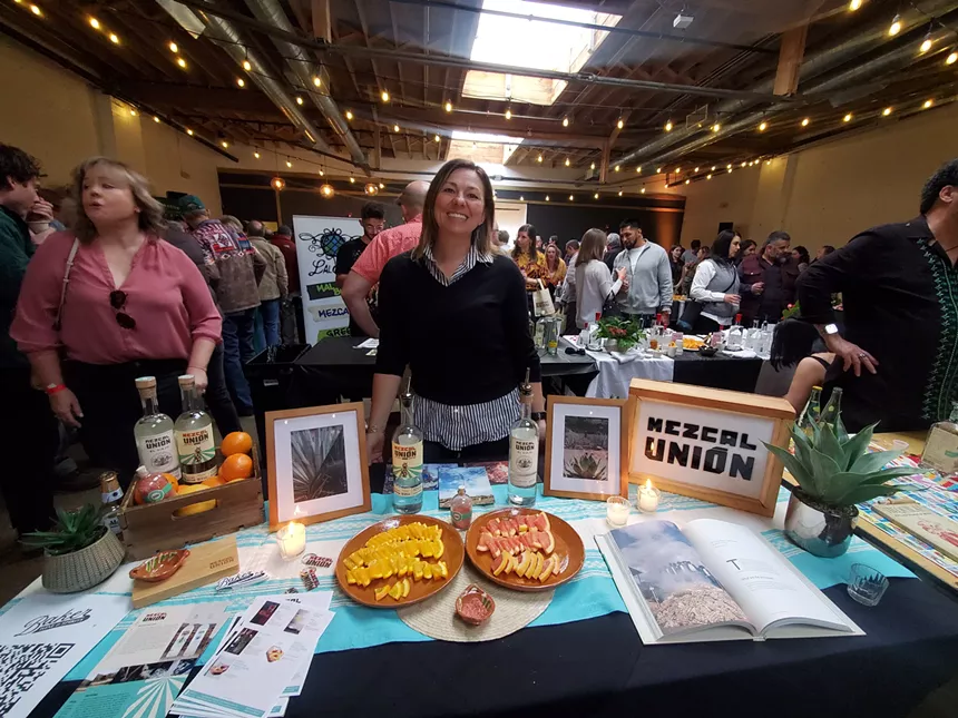 a woman standing behind a table with bottles of mezcal and sliced fruit