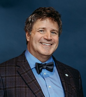 A man wearing a suit jacket and bowtie smiles at the camera