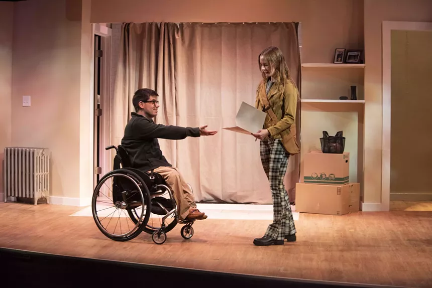 person in a wheelchair on stage reaches for a folder from a woman in plaid pants