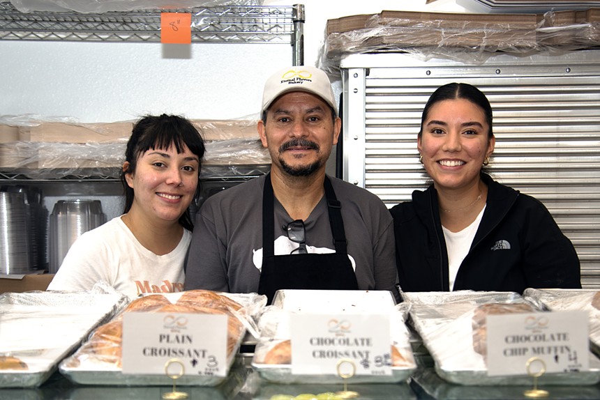 three people posing behind a counter with pastries on it