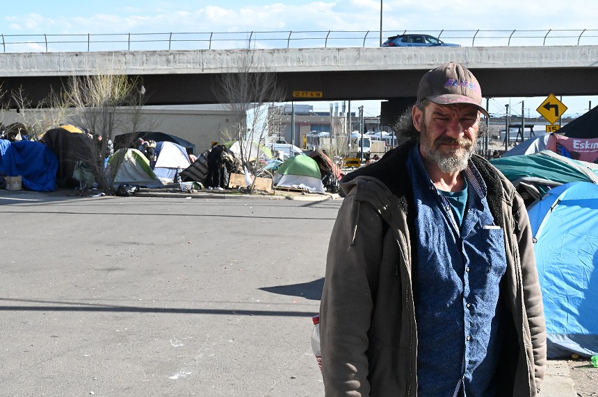 A homeless man stands in front of an encampment.