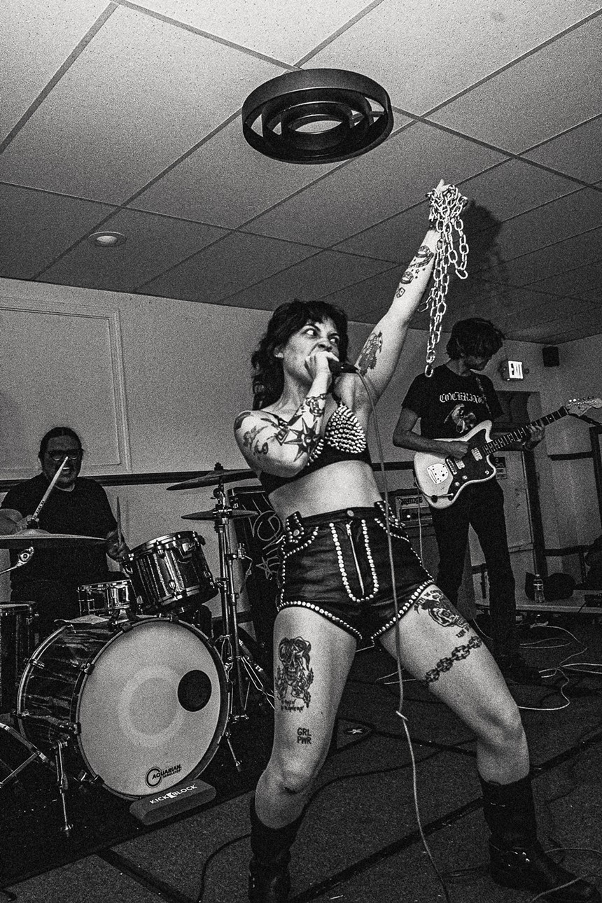 woman wearing studded leather sings into a mic while holding a chain