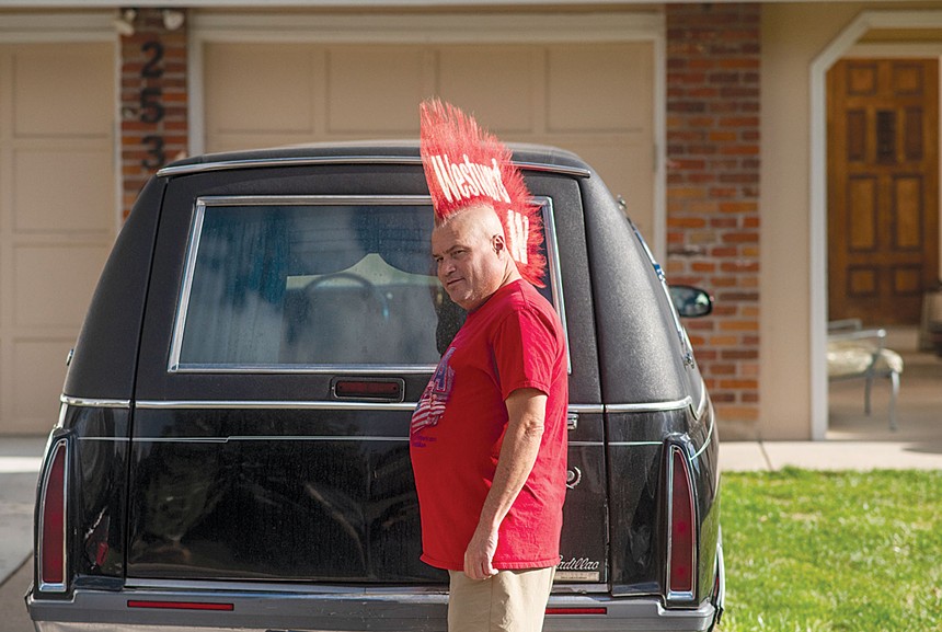 man in red shirt with red mohawk stands in front of a hearse