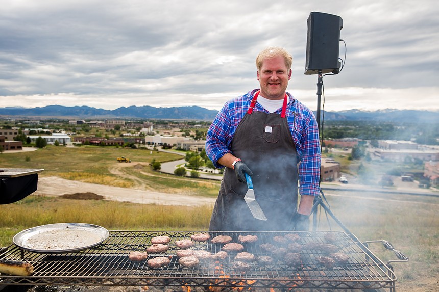 a man in an apron holding a spatula in front of a large grill outside with mountains in the background