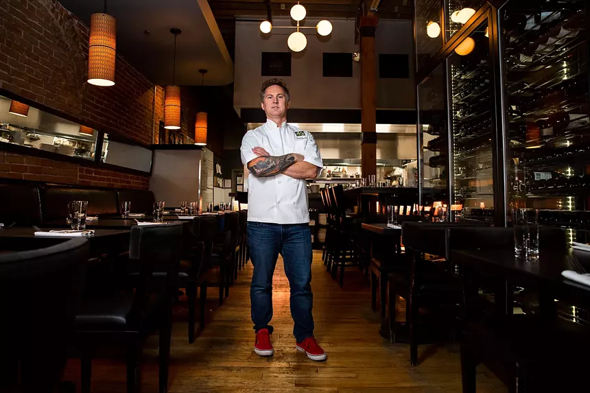 man in a white chef coat standing in a dining room with his arms crossed