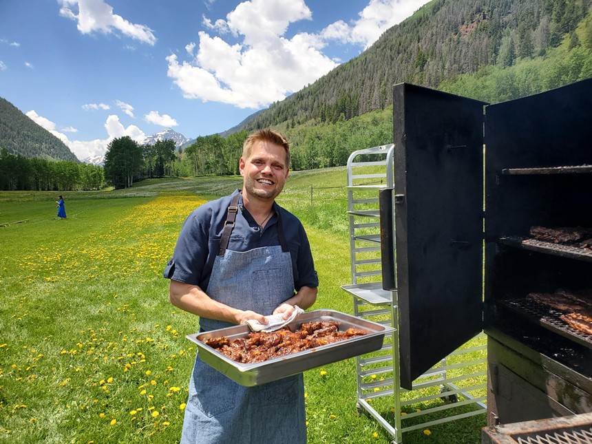 man in an apron holding a pan of chicken in a field with a mountain backdrop