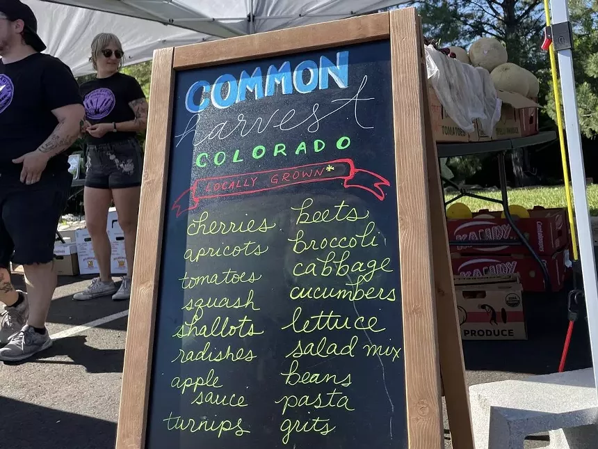 A chalkboard sign next to the Common Harvest Colorado farmers market booth listing available produce and pantry items