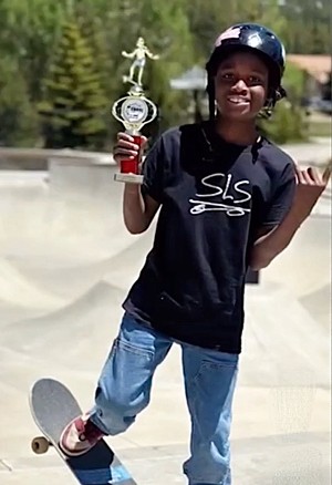 An 11-year-old boy smiles at a skate park, holding a first-place trophy and standing on a skateboard.