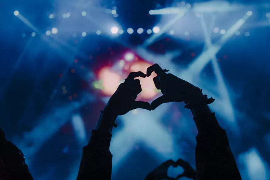 A photo showing someone making a heart gesture with their fingers while at a concert.