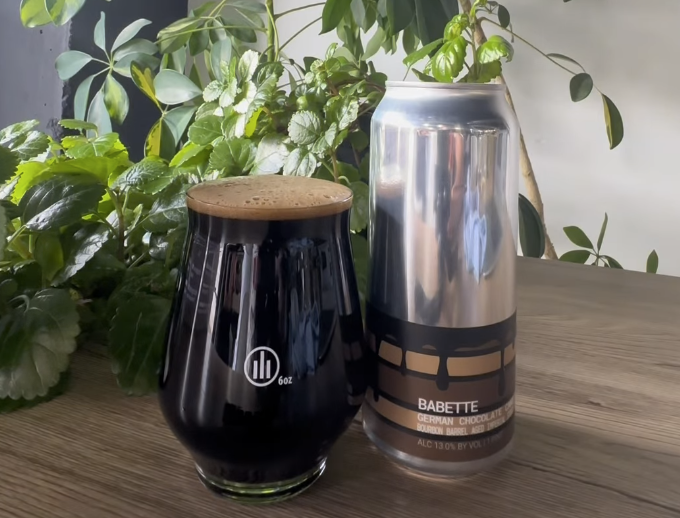 Can of beer with dark contents in a glass, background greenery.