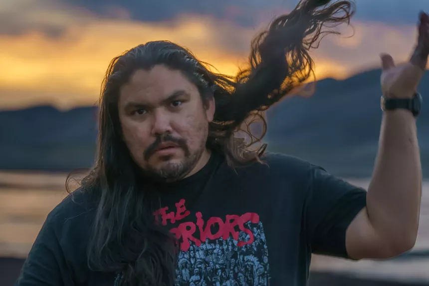 A man with long hair flips it back as the sun sets in the distance.